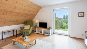 Een moderne woonkamer met een schuine kap, een groene fauteuil, een houten salontafel, een televisie op een witte dressoir en een groot raam met uitzicht op een bos.
