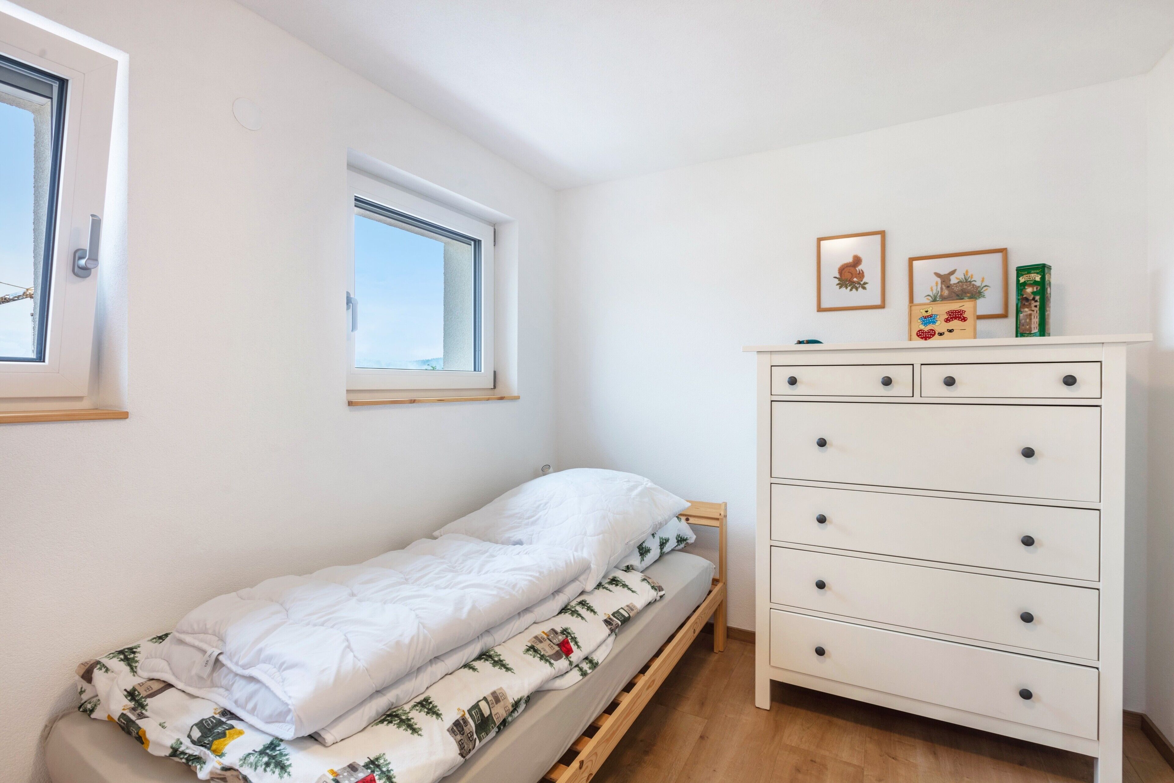 Ein kleines Zimmer mit einem Einzelbett, einem Holzrahmen, einer weißen Kommode mit Schubladen und einem Fenster mit Blick zum Himmel.