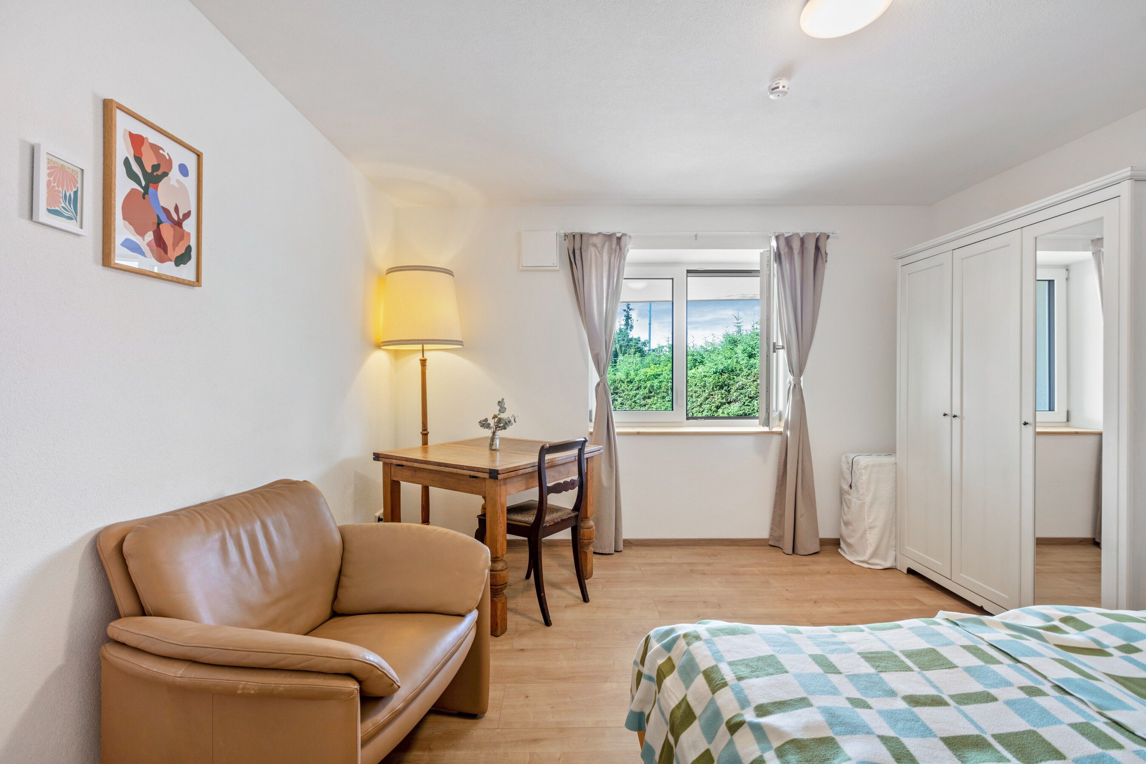 A bedroom with a bed, a sofa, a table with a lamp, a window with curtains, and a wardrobe.