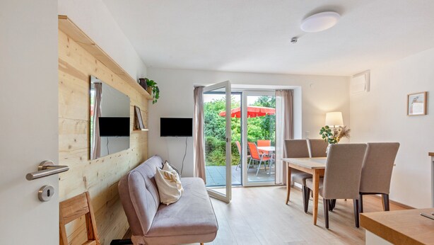 Een moderne woonkamer met een bank, een eettafel met stoelen, een aan de muur bevestigde televisie en een schuifdeur naar een buitenruimte met een rode parasol.