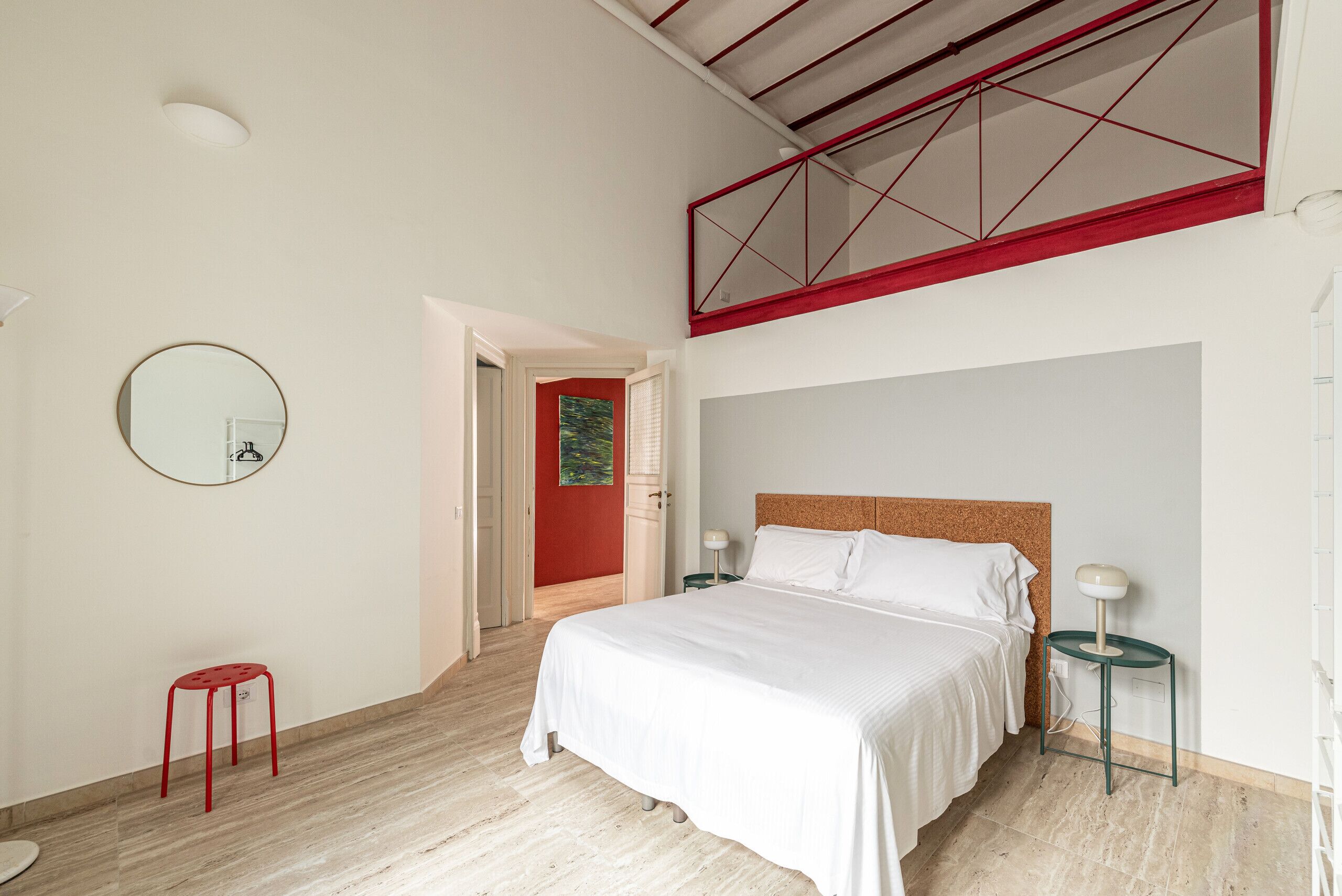 Un dormitorio con cama, un taburete rojo, un espejo redondo y una puerta que da a otra habitación.