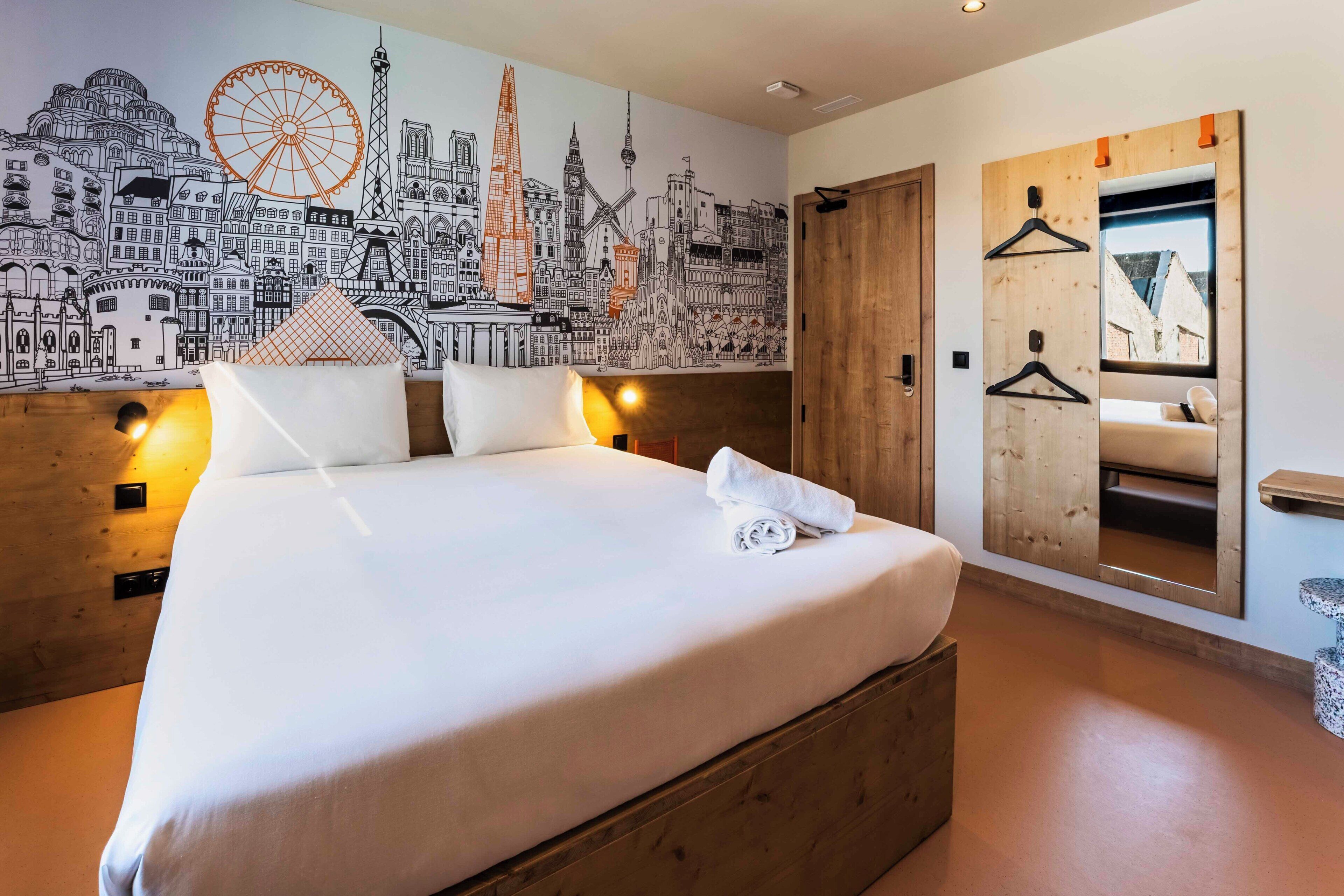 Habitación de hotel con una cama grande, un mural de paisaje urbano, cabecero de madera y un espejo con puerta de madera.