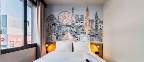 Una habitación de hotel con una cama, un mural de paisaje urbano en la pared, una ventana con cortinas y vistas a un edificio con un cartel que dice 'deluxe'.