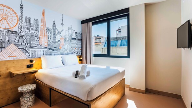 A hotel room with a bed, a small round table, a wall mural, a window with a view, and a flat-screen TV. - easyHotel Madrid Alcala (Madrid)