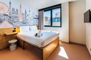 A hotel room with a bed, a small round table, a wall mural, a window with a view, and a flat-screen TV. - easyHotel Madrid Alcala (Madrid)