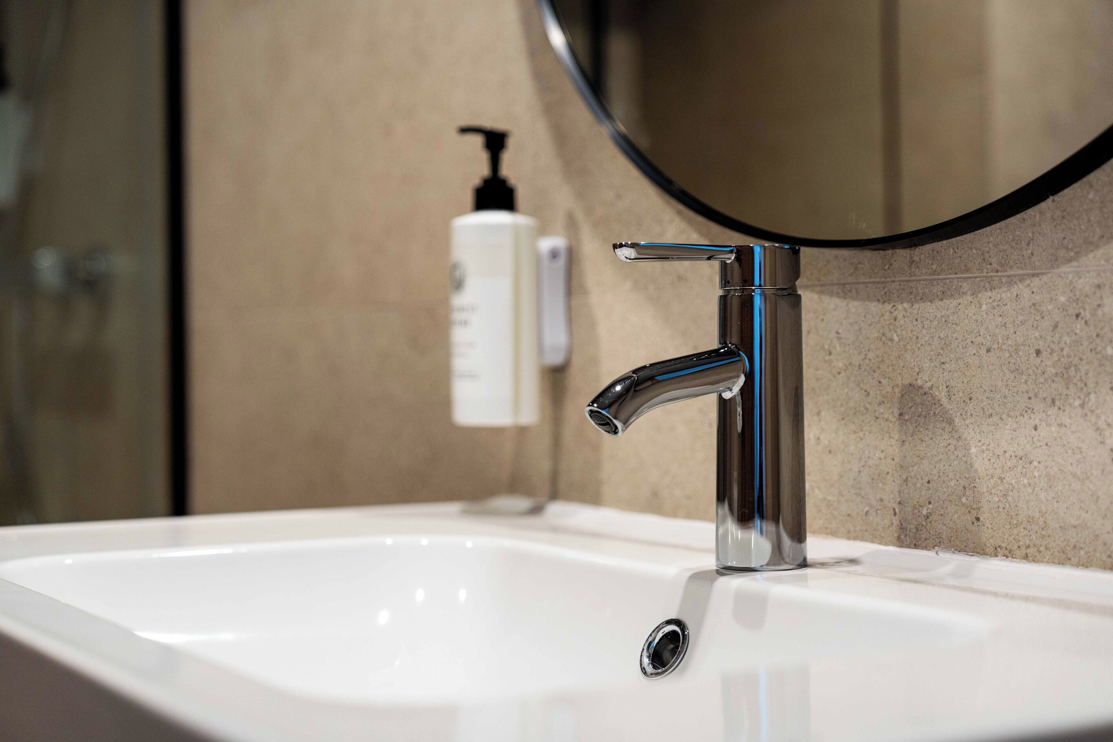 Ein modernes Waschbecken mit chromfarbenem Wasserhahn, einem Spiegel und einer Handseifenflasche.