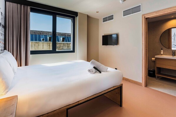 Habitación de hotel con cama, televisor, espejo y ventana con vista a edificios.