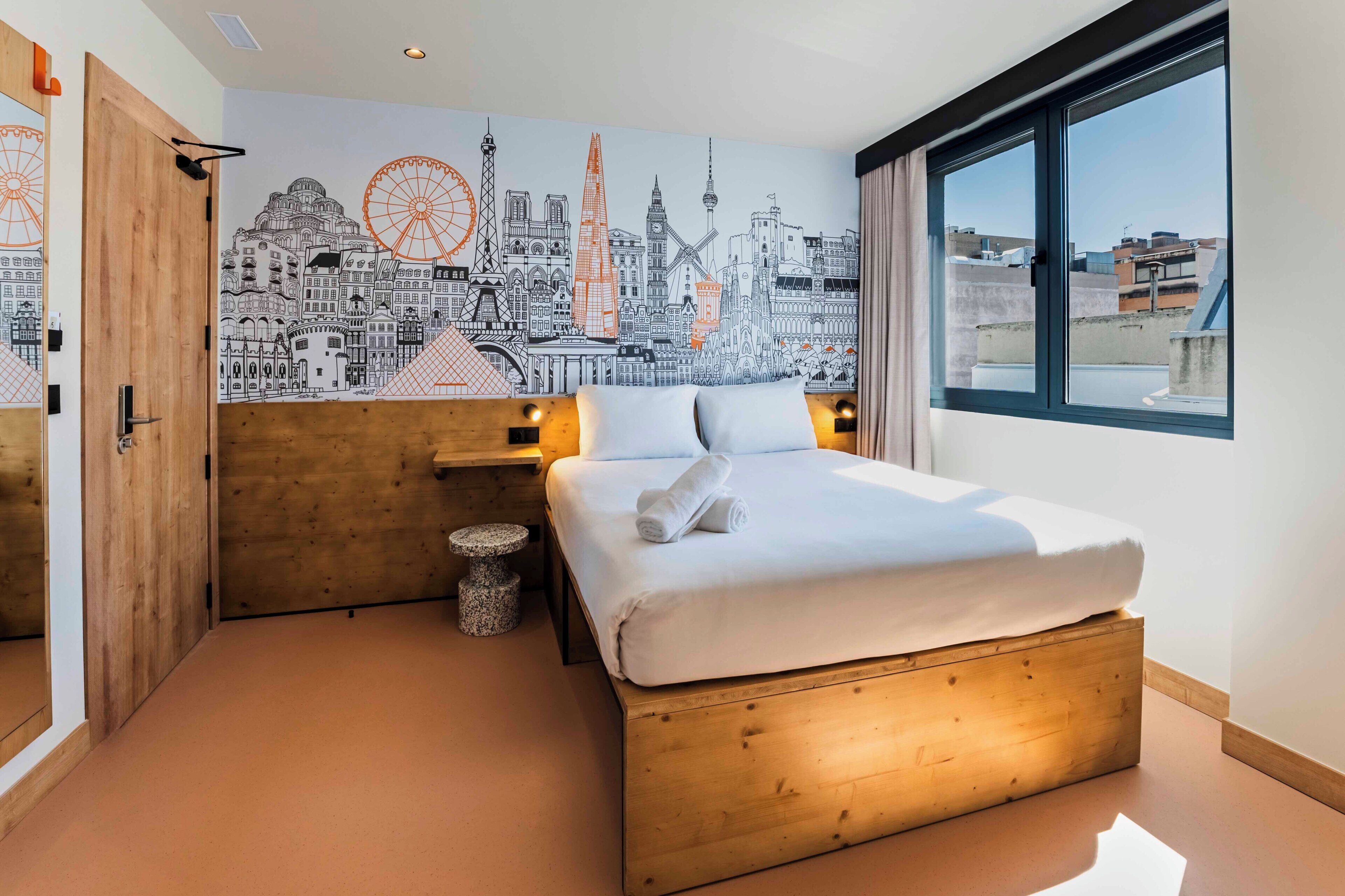 A bedroom with a cityscape mural, a wooden bed, bedside lamps, and a window with a view.