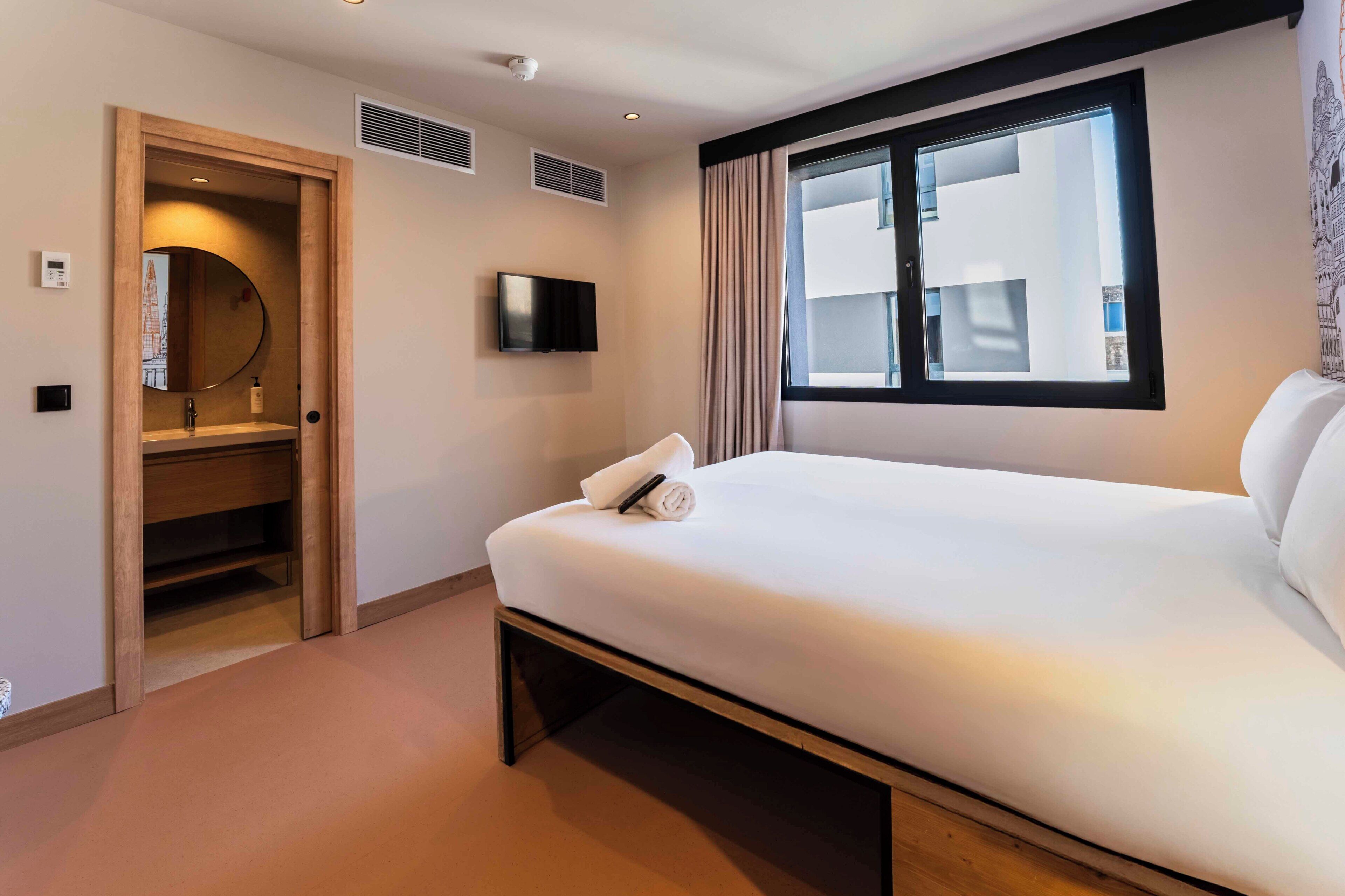 A hotel room with a bed, a television, a window with curtains, a door leading to a closet, and a wall-mounted air vent.