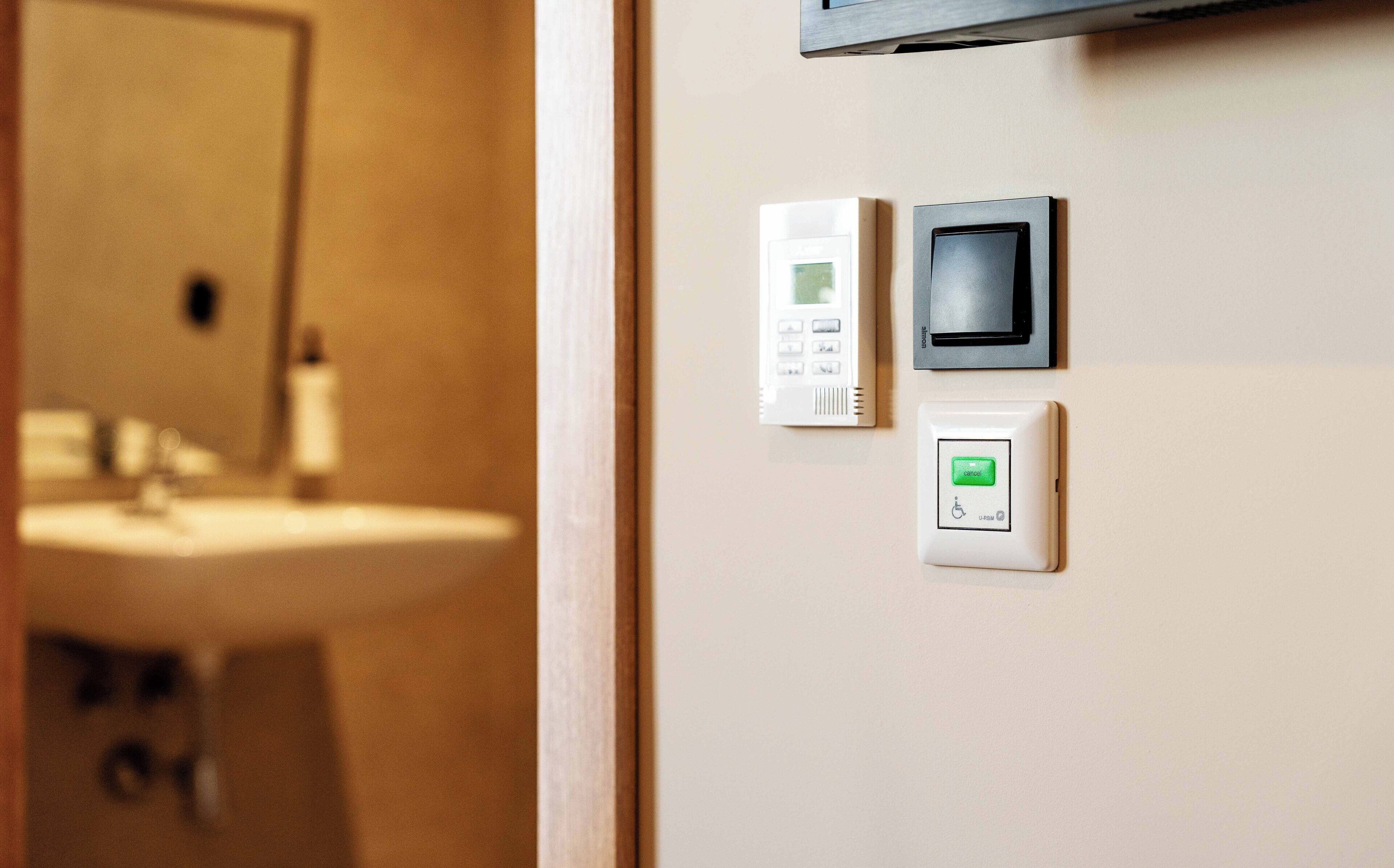 A bathroom with a sink, a mirror, and a wall-mounted control panel with a green button and a wheelchair accessibility symbol.