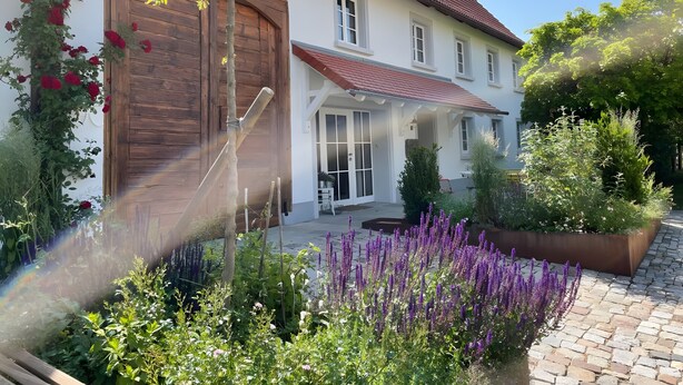 A two-story house with a large wooden garage door, a covered porch, and a garden with various plants and flowers. - Apartment 'Ferienwohnung' with Private Garden and Terrace, Wi-Fi Included (Hüfingen)