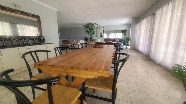 Un comedor espacioso con una larga mesa de madera y sillas negras, rodeado por una cocina y una sala de estar con sofá y plantas.