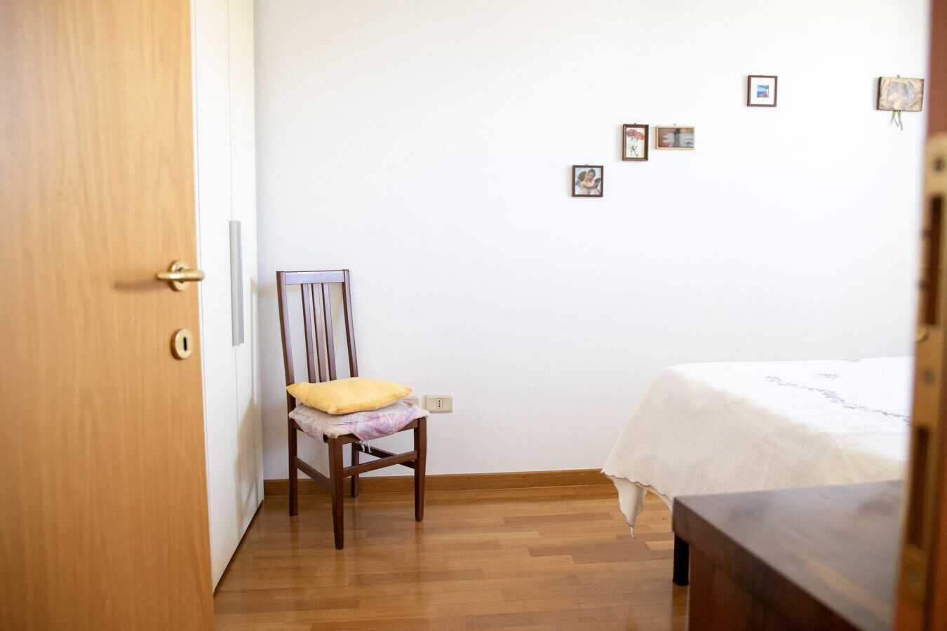 Um quarto com porta de madeira, uma cadeira de madeira com almofada, uma cama com colcha branca e uma parede com várias fotos emolduradas.