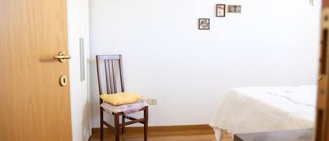 Um quarto com porta de madeira, uma cadeira de madeira com almofada, uma cama com colcha branca e uma parede com várias fotos emolduradas.
