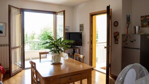 Een eethoek met een houten tafel, stoelen, een plant in pot, een televisie en een koelkast.