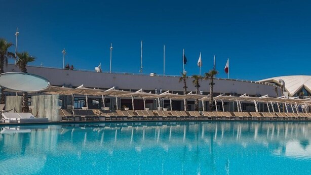 En stor swimmingpool med liggestole og parasoller, omgivet af en bygning med flag og palmer.