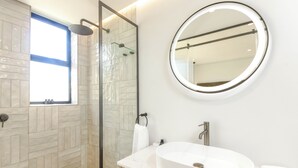 Un baño moderno con ducha envuelta en vidrio, un espejo redondo y un lavabo blanco.