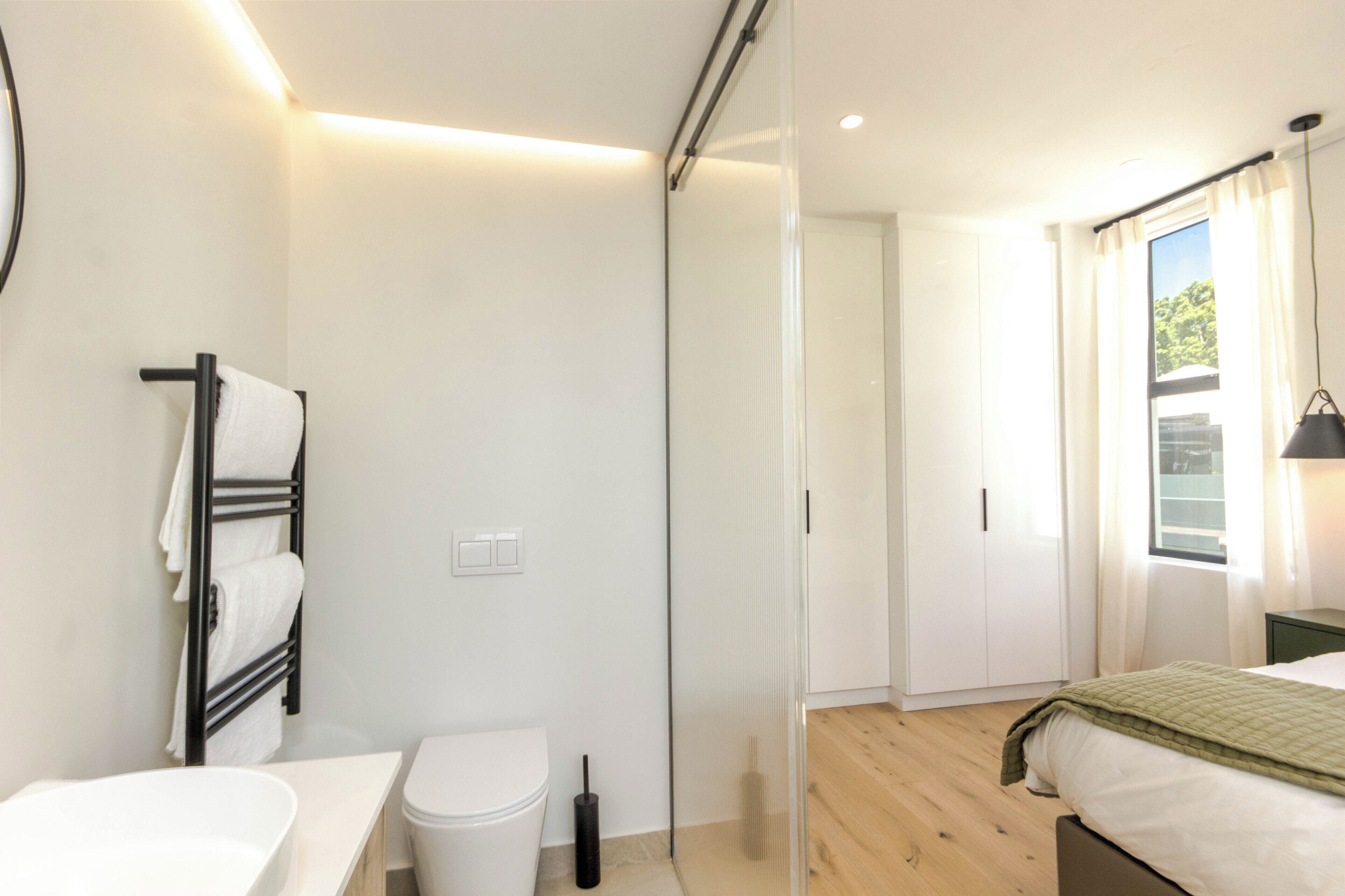 Un dormitori modern amb terra de fusta, un llit amb llençol verd, un vàter blanc, una cabina de banyera de vidre i un bany amb un lavabo blanc i porta tovalloles.