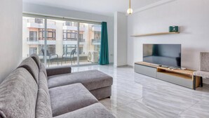 Ein modernes Wohnzimmer mit einer grauen Couch, einem Flachbildfernseher und einem Balkon mit Blick auf andere Gebäude.