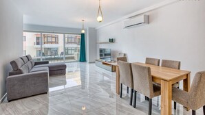 Una sala de estar moderna con un conjunto de sofás, una mesa de comedor con sillas y un televisor. Hay un ventanal que ofrece vistas al exterior.