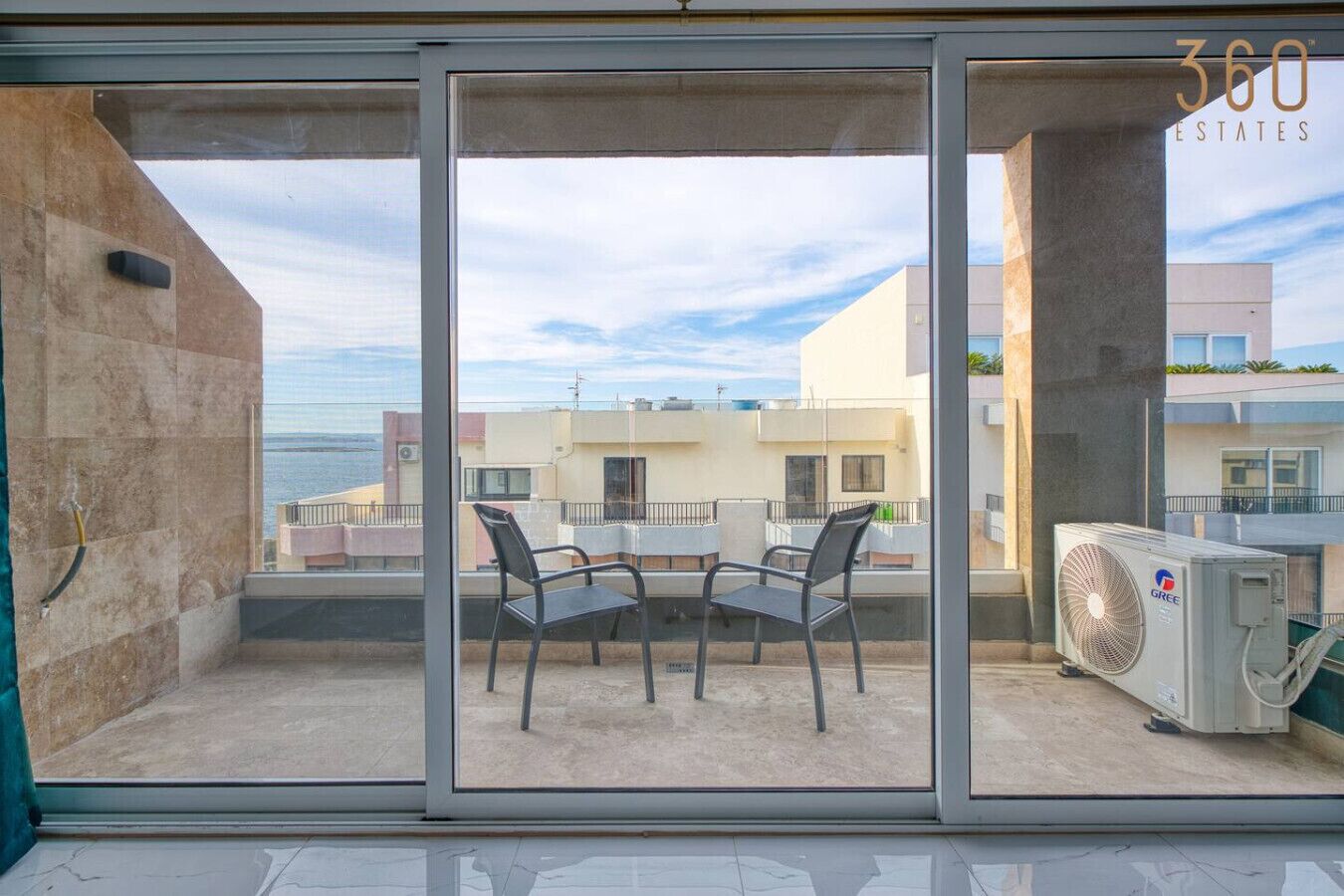 A balcony with a table and chairs, a fan, and a view of buildings and the sea.