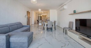 A modern living room with a grey sofa, a TV on a wooden stand, and a dining area with chairs and a table. - A 2BR designer seafront home w/Wifi, AC and Beach (St. Paul`s Bay)