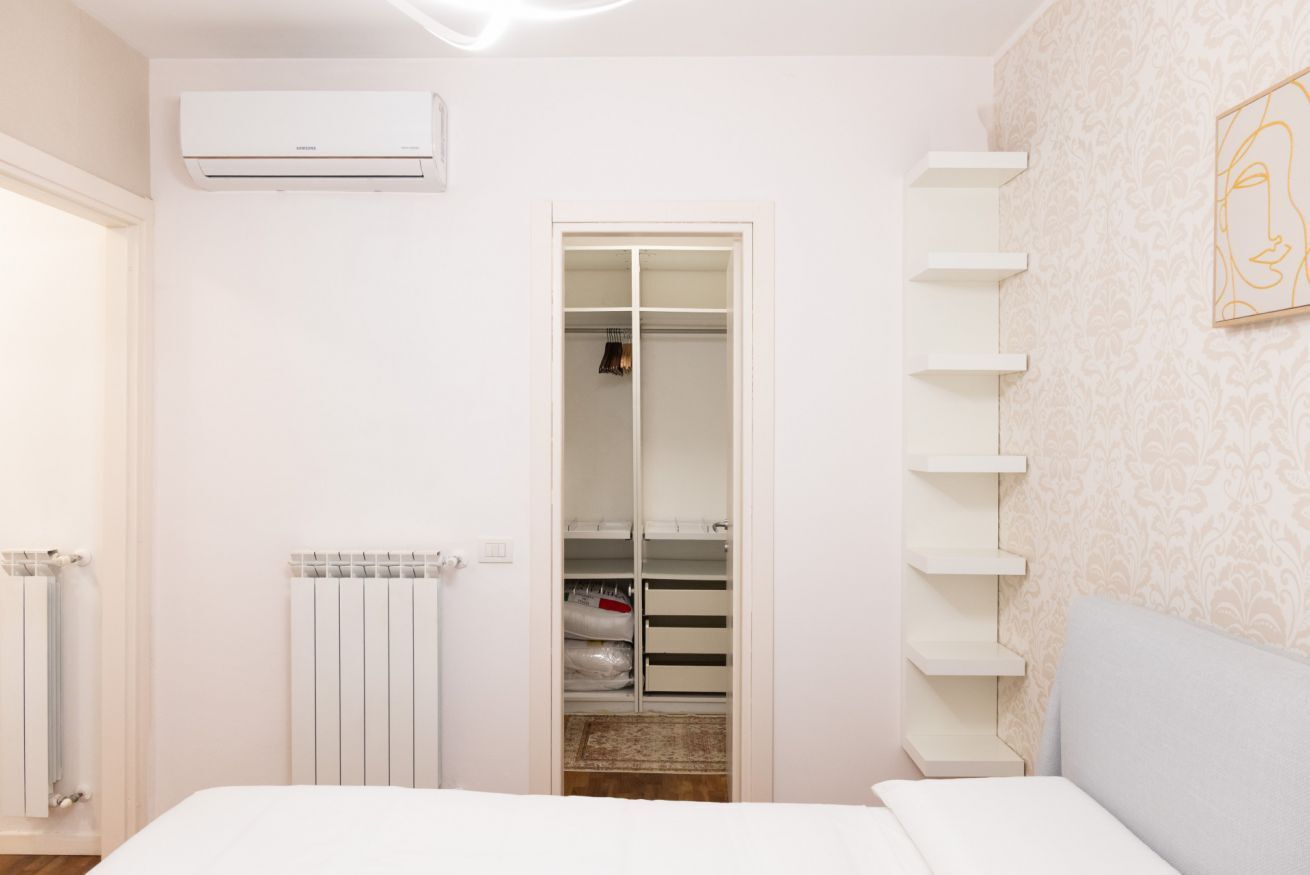 A bedroom with a bed, a white radiator, an air conditioner, a closet with shelves, and a patterned wall.