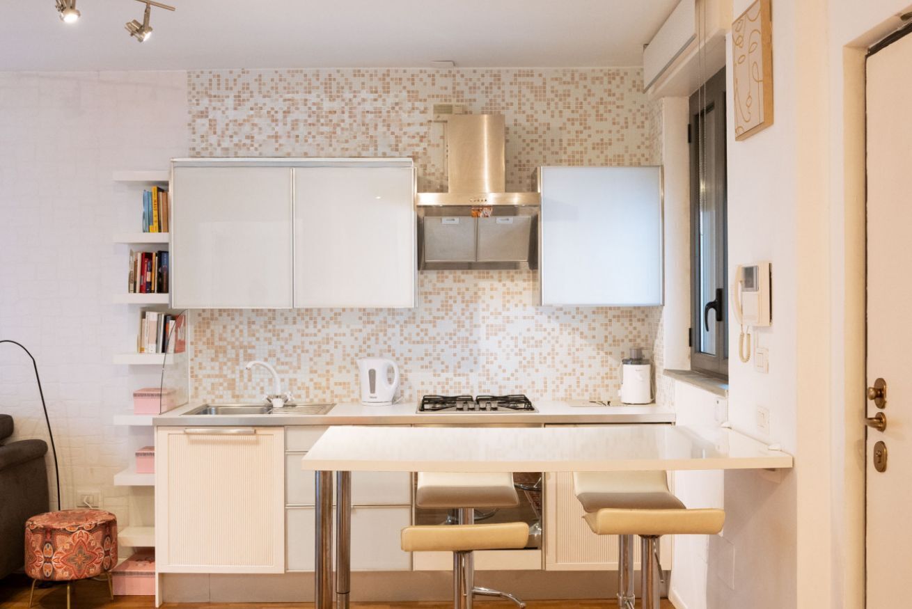 Une cuisine compacte avec des meubles blancs, une cuisinière à gaz et un petit évier. Il y a deux tabourets au comptoir et un tabouret à motifs dans le coin.