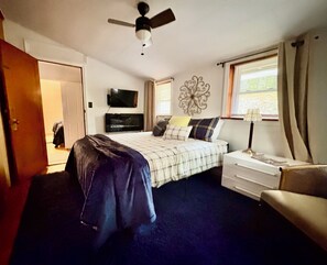 Slaapkamer met een bed, een televisie, een plafondventilator, een raam met gordijnen en een nachtkastje met een lamp.
