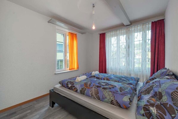A bedroom with two beds, a window with orange curtains, a window with red curtains, and a ceiling light.