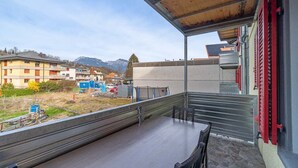 Un balcon avec une table et des chaises, donnant sur un parking et des immeubles aux volets rouges.