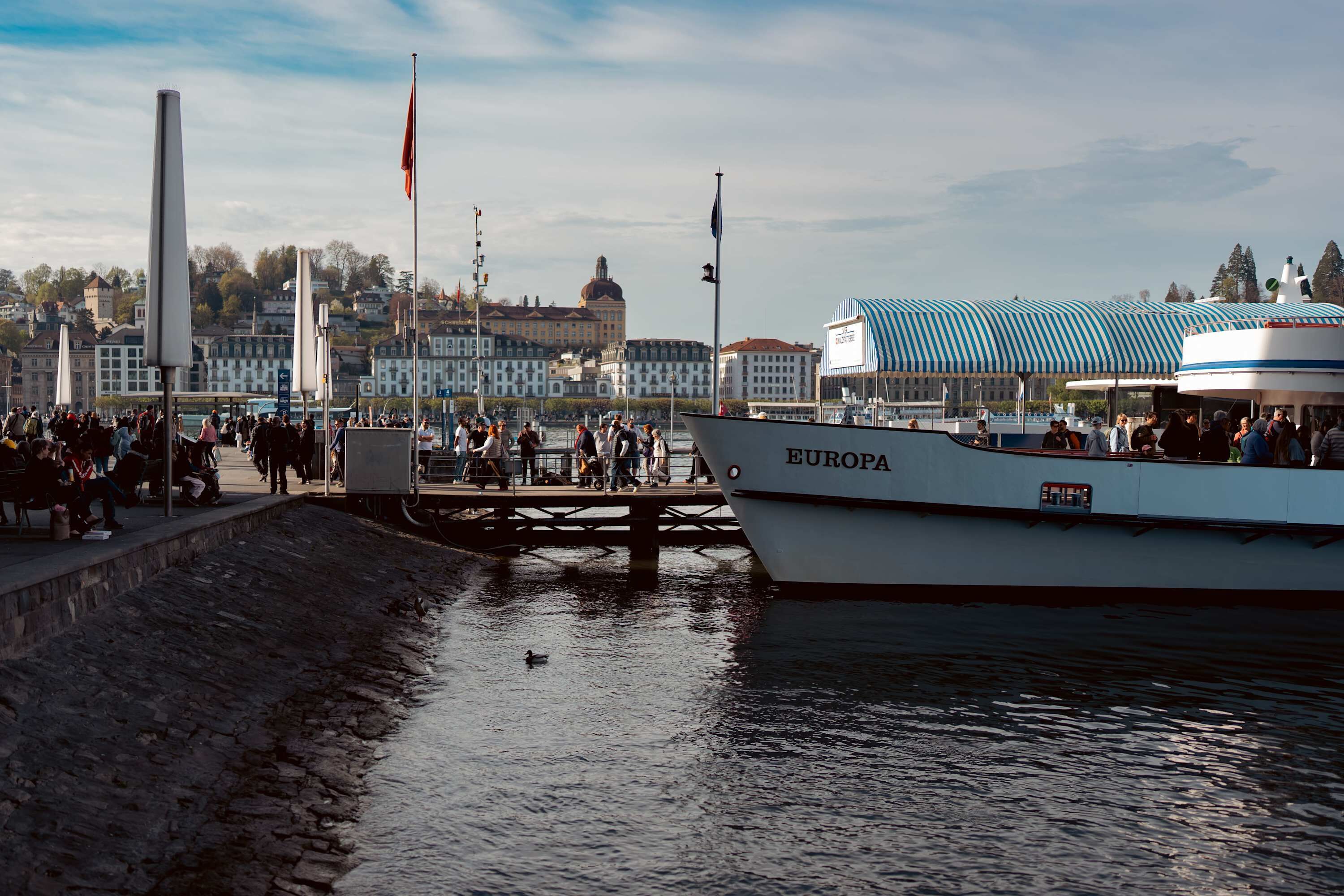A waterfront area with a docked boat named 'EUROPA,' a stone embankment, and a cityscape in the background.