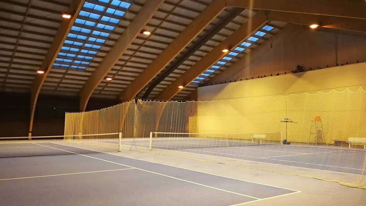 An indoor tennis court with a net, enclosed by walls and a roof with skylights.