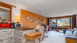 Une cuisine moderne avec un mur en bois, un plan de travail rouge et une table en bois. Le salon comprend un canapé et un fauteuil, et offre une vue sur un paysage montagneux.