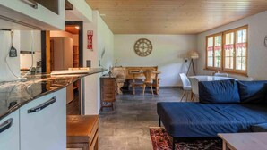 A modern kitchen with a wooden ceiling, a dining area with a wooden table and chairs, and a blue sofa in the living area. - With Hot Tub, Sauna, Garden|with View near Gondola (Diemtigen)