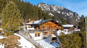 A log cabin with a balcony, surrounded by snow and trees, with a mountain in the background.