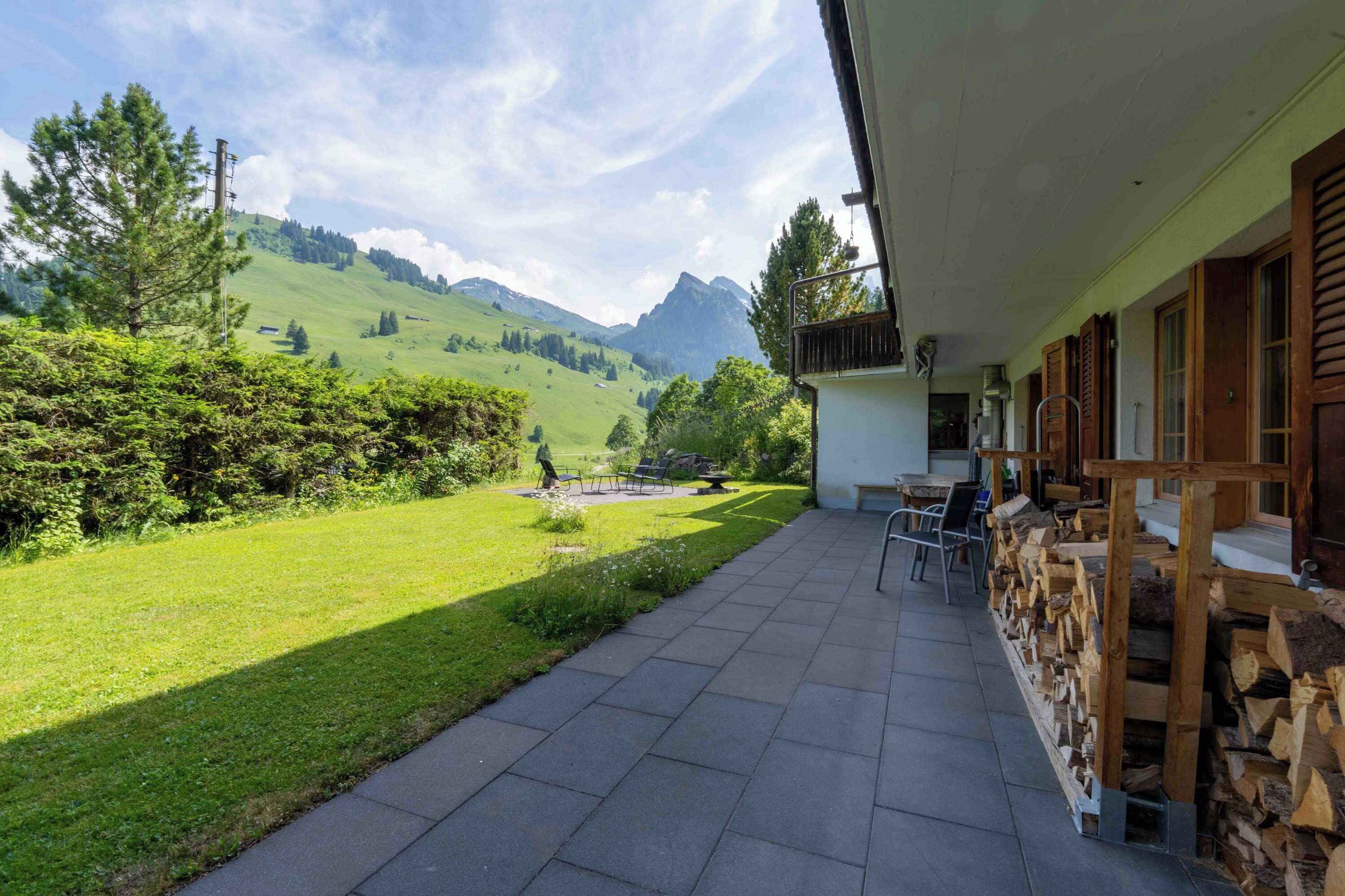 A patio with a view of green hills and mountains, a wooden fence, and a stack of firewood.