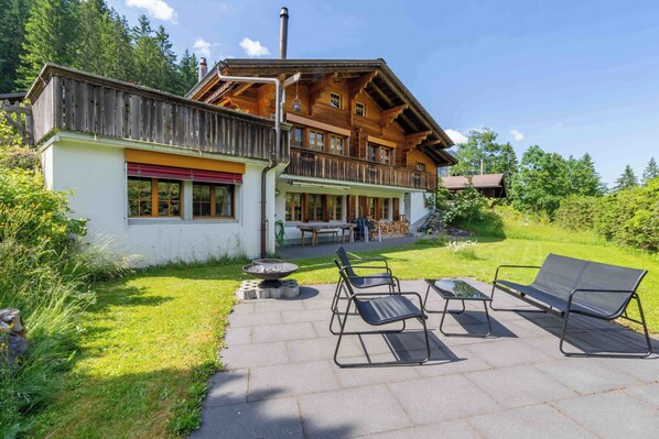 A chalet with a wooden balcony, a patio with outdoor furniture, and a view of the surrounding forest.