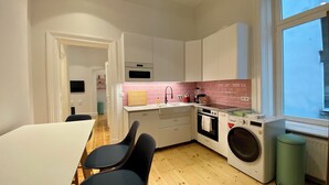 A compact kitchen with white cabinets, a pink-tiled backsplash, a washing machine, and a window with a view. - STACEY Downtown (Hamburg)