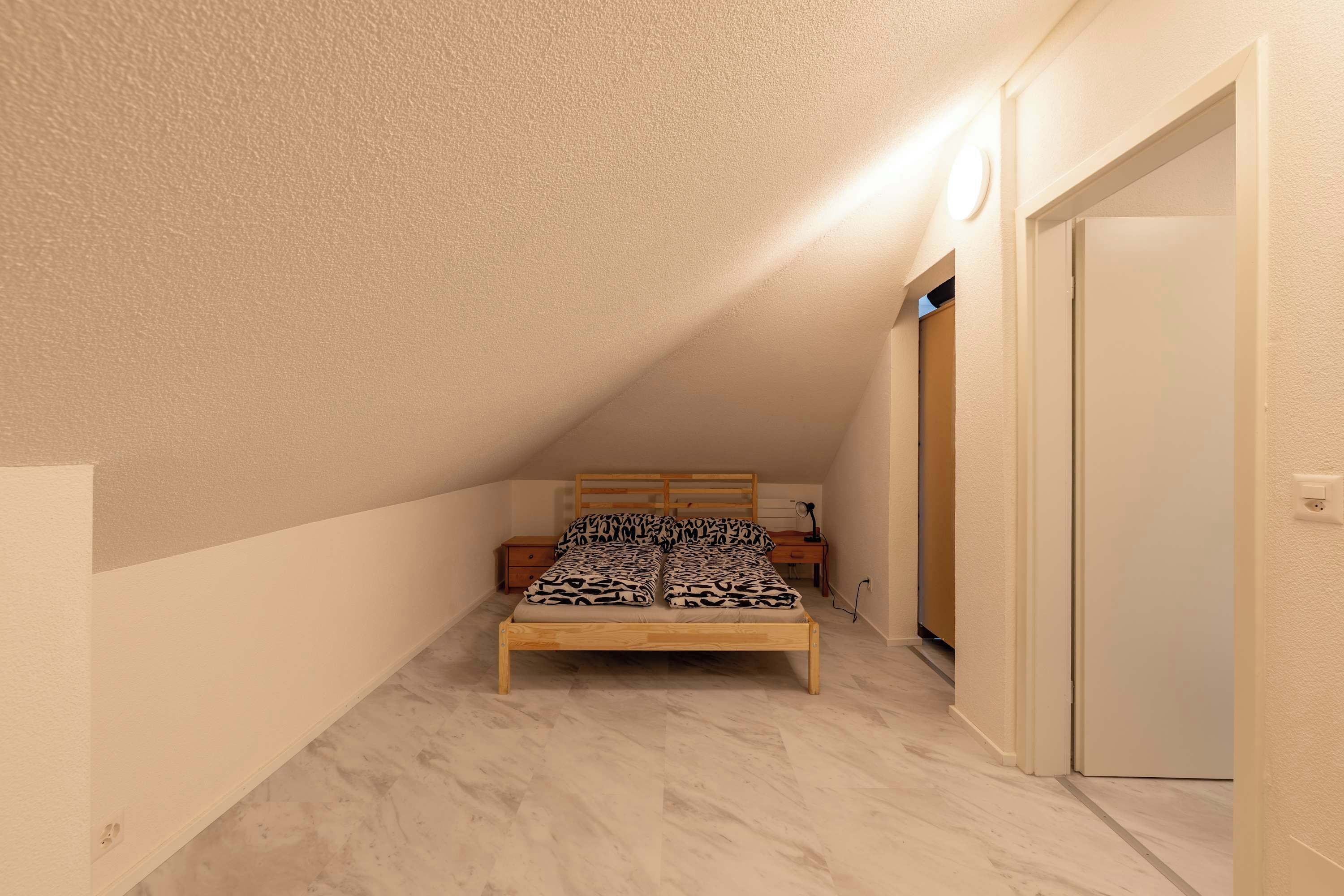 A small, narrow room with a sloped ceiling, a single bed with patterned bedding, a nightstand, and a door.