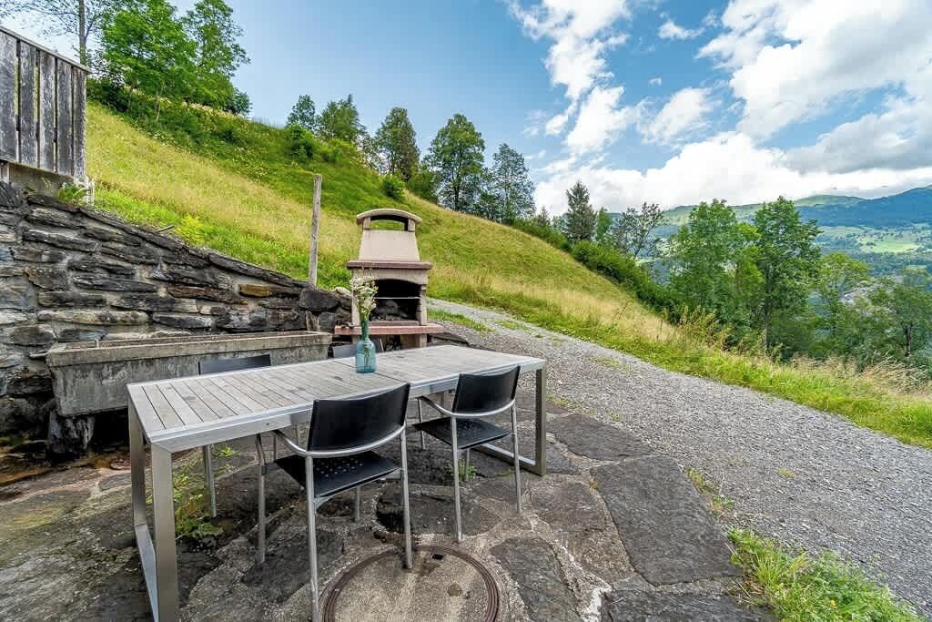 Ein Essbereich im Freien mit einem Steintisch und -stühlen, einer Steinmauer und Blick auf einen grasbewachsenen Hügel und Bäume.