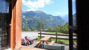 Vista desde una habitación con ventana a un paisaje montañoso, un banco de madera y un coche de juguete rojo.