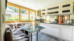Eine moderne Küche mit weißen Schränken, einem Glastisch und Blick auf ein Haus durch große Fenster.
