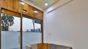 A modern room with a wooden ceiling, a sliding glass door leading to an outdoor area, and a grey sofa. - Hotel Krishna Palace (Becharaji)