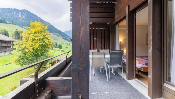Ein Balkon mit Blick auf ein grünes Tal und Berge, ein Tisch mit Stühlen und ein Bett im angrenzenden Zimmer.