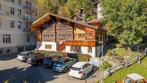 A multi-story building with a wooden facade, a parking area with several cars, and a mountainous backdrop. - Mountain View Studio Adelboden|Gondola & Spa (Adelboden)