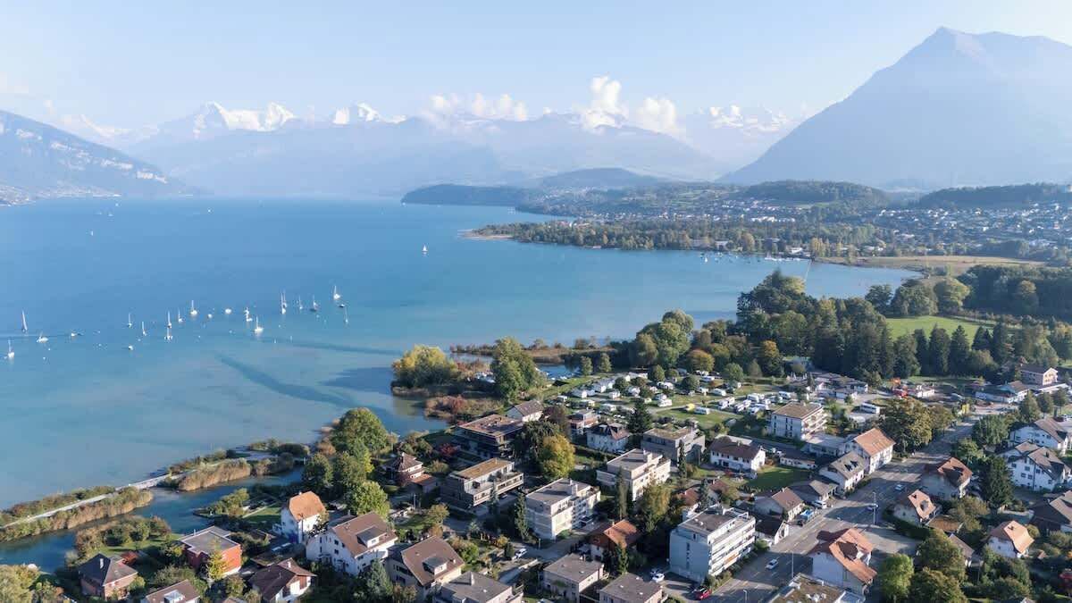 Une vue aérienne d’une ville située au bord d’un lac, avec des immeubles résidentiels, des bateaux sur le lac et des montagnes en arrière-plan.