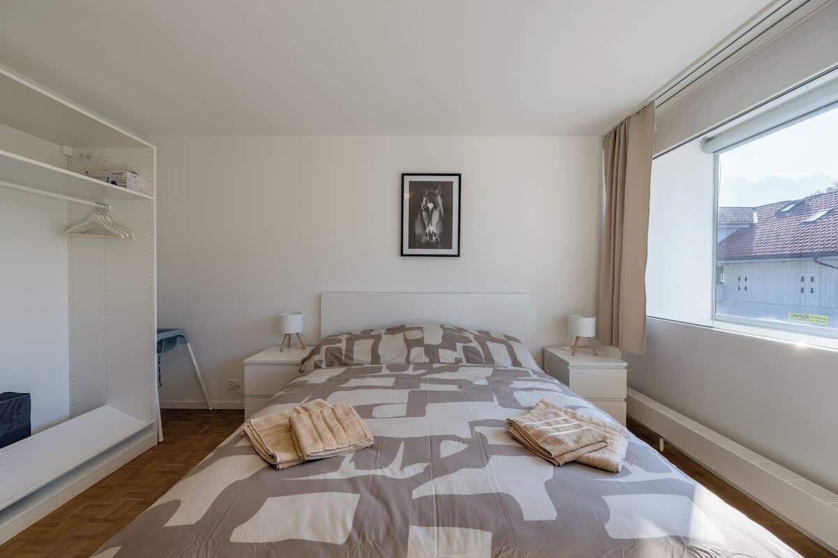 Un dormitorio con cama, mesita de noche, armario y ventana con vistas a edificios.