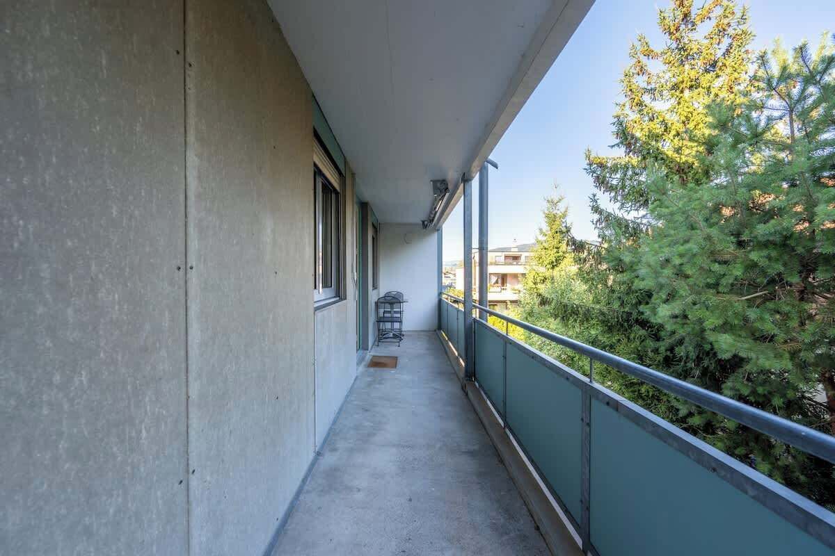 Ein schmaler Balkon mit Betonboden, Metallgeländer und Blick auf Bäume und Gebäude im Hintergrund.