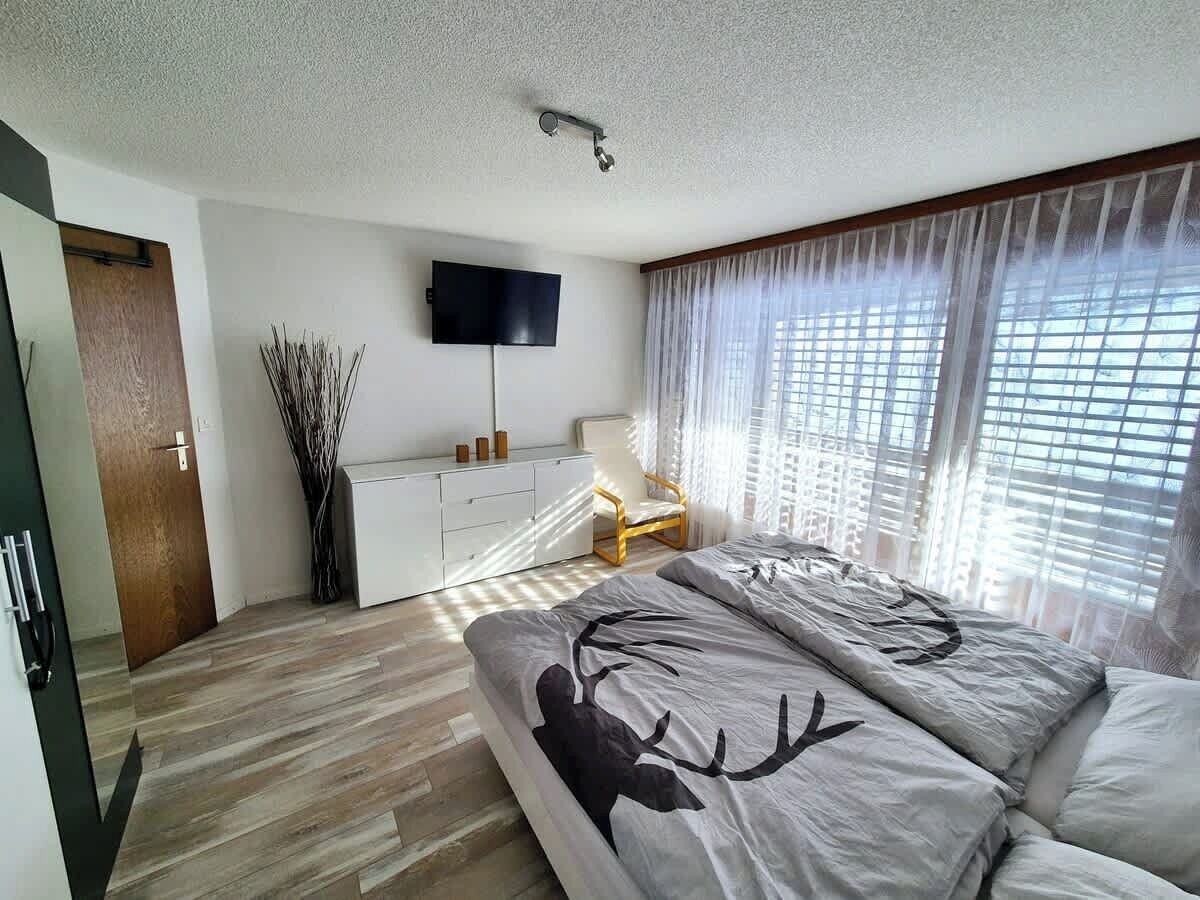 Une chambre à coucher avec un lit, un téléviseur fixé au mur, une fenêtre avec des rideaux légers et un plancher en bois.